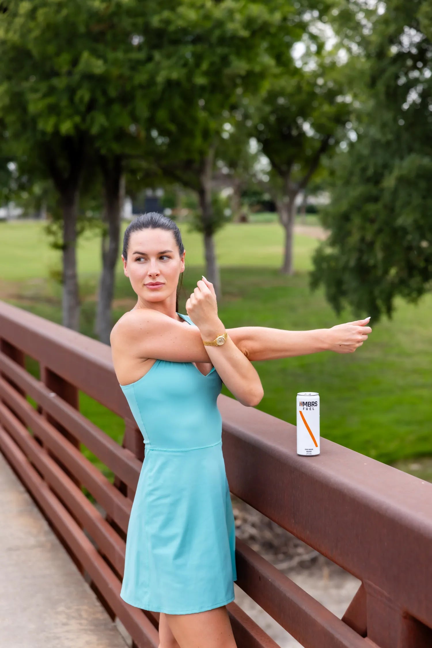 Woman in a blue dress stretching outdoors on a bridge with a can of NMBRS Fuel in the background.