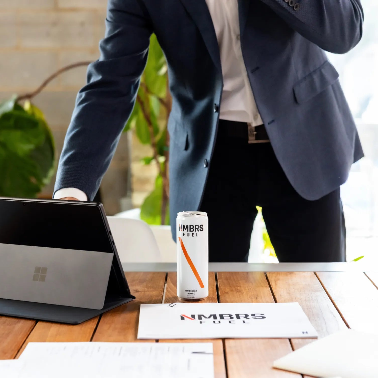 Person in a suit standing next to a table with a tablet and a can of NMBRS Fuel Energy drink.