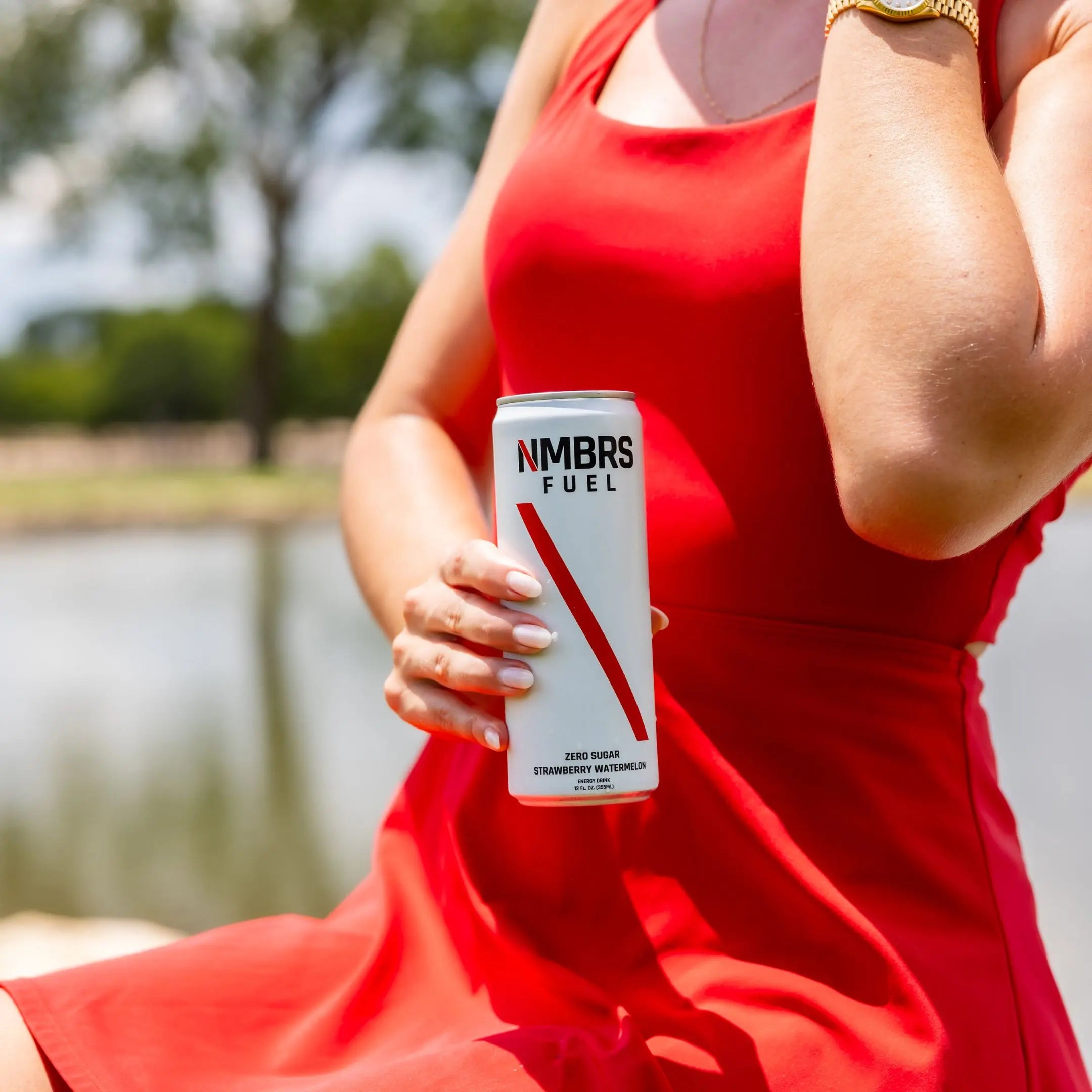 Women in a red dress holding a can of NMBRS FUEL by a lake.