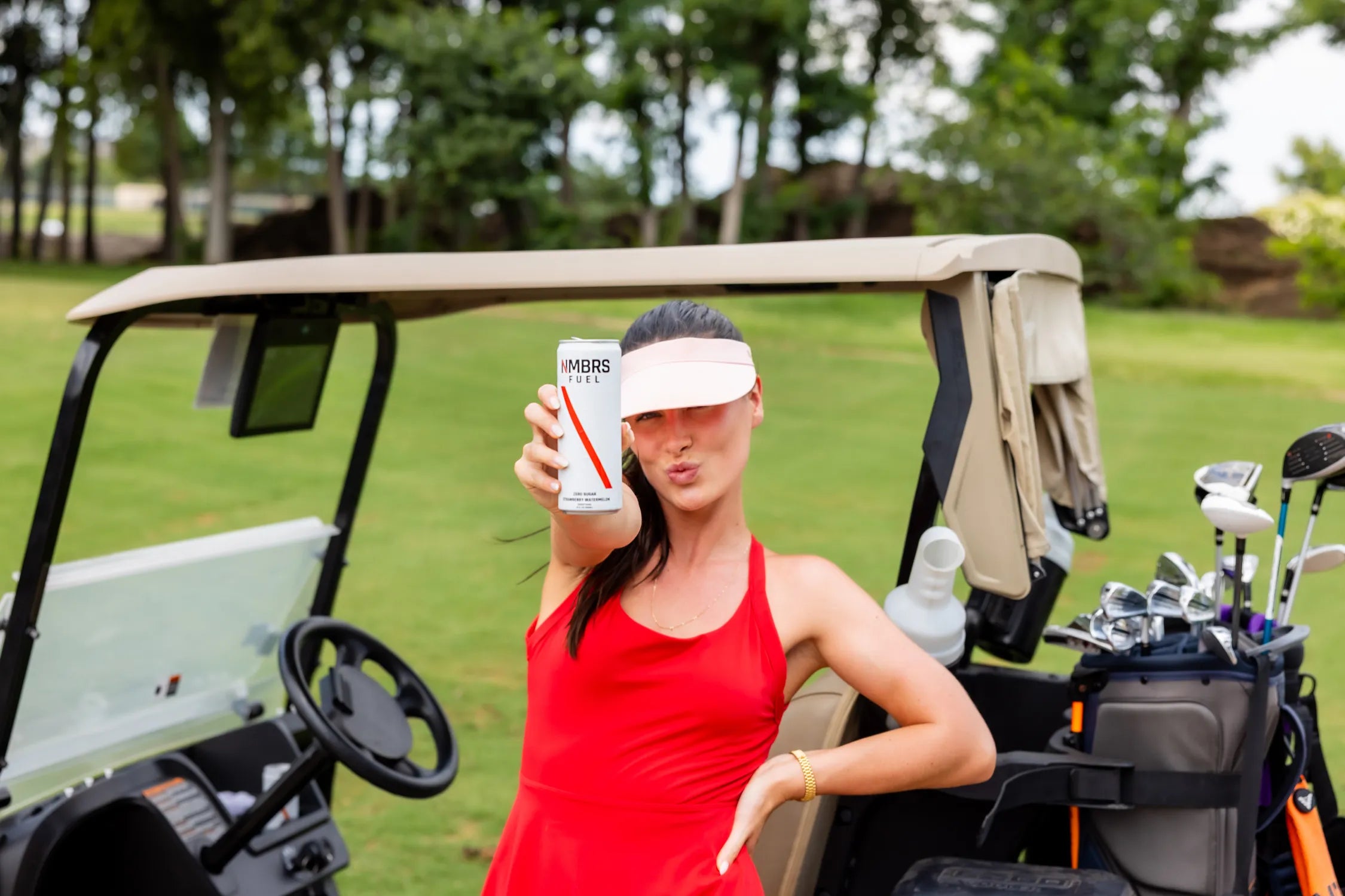 Woman in a red dress holding a can of NMBRS Fuel next to a golf cart on a golf course